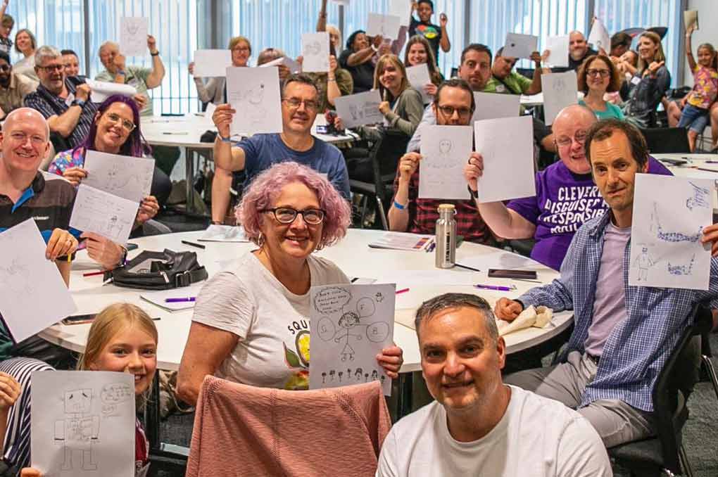A conference room full of people holding up pieces of paper with cartoons they drew on them.